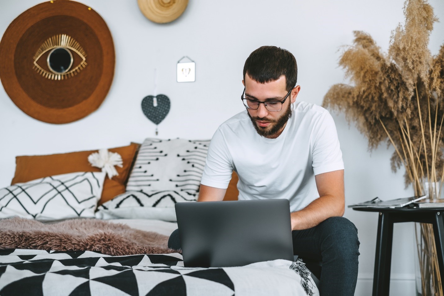 A person sitting on a bed is looking at a laptop. The room is well decorated with soft gray, white, and brown hues.