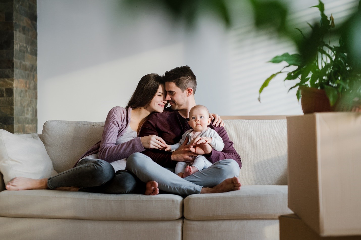A young couple and baby smiling together on a couch in their new home, surrounded by boxes and plants.