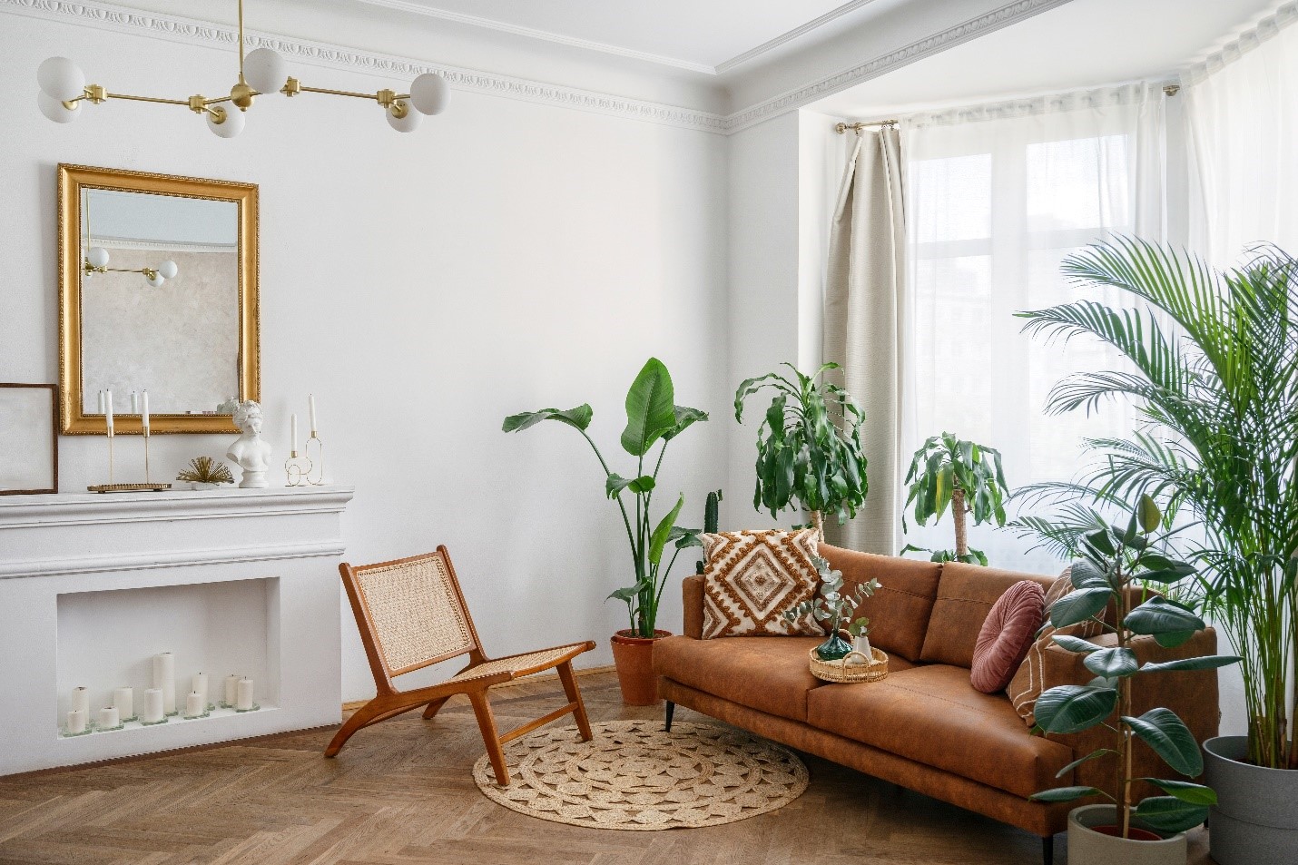 A sitting room interior with natural light filling the space. There is a brown couch and a multitude of plants. Perpendicular to the window, a gold framed mirror is over a white, decorated hearth.