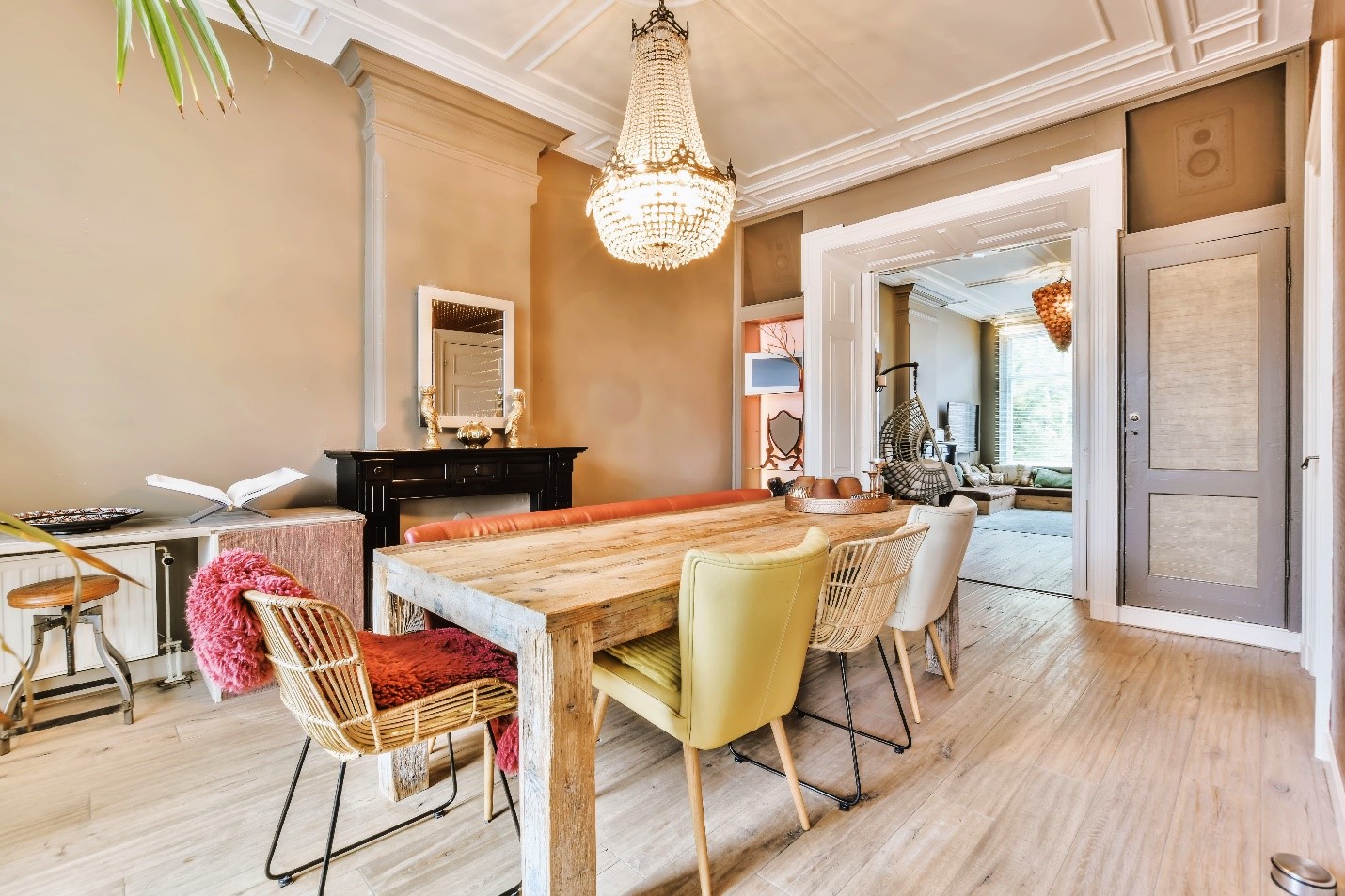 A large dining room with a light wood dining table and mismatched chairs. There is a brilliant chandelier above. The room's door frame, wall trim, and ceiling are painted white.