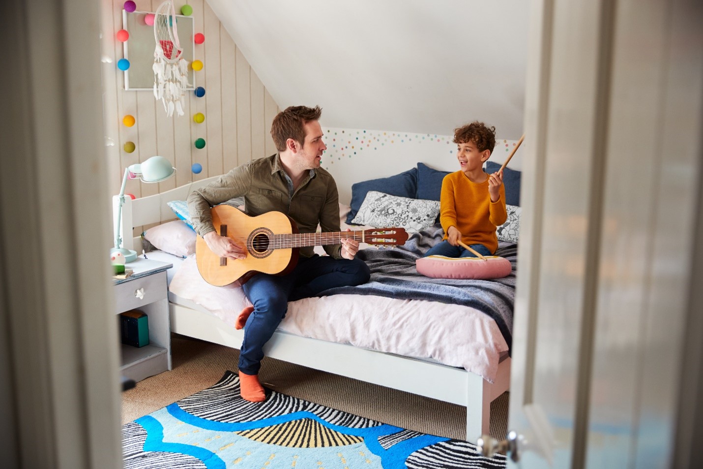 A parent playing guitar and singing with their child who is playing drums on a pillow.