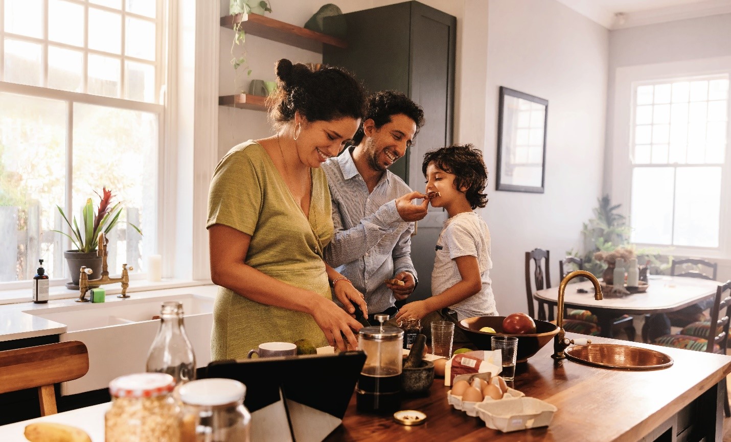 A family cooking together in their kitchen. A boy sits on the kitchen counter, being fed a small bite of bread from his dad. The mother stands next to them with a smile, cutting up vegetables.