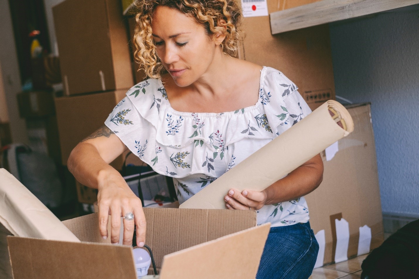 A woman sits on the ground with boxes around her. She's leaning over an open box and holding a wrapped poster. Her other arm is entering the box to rummage.