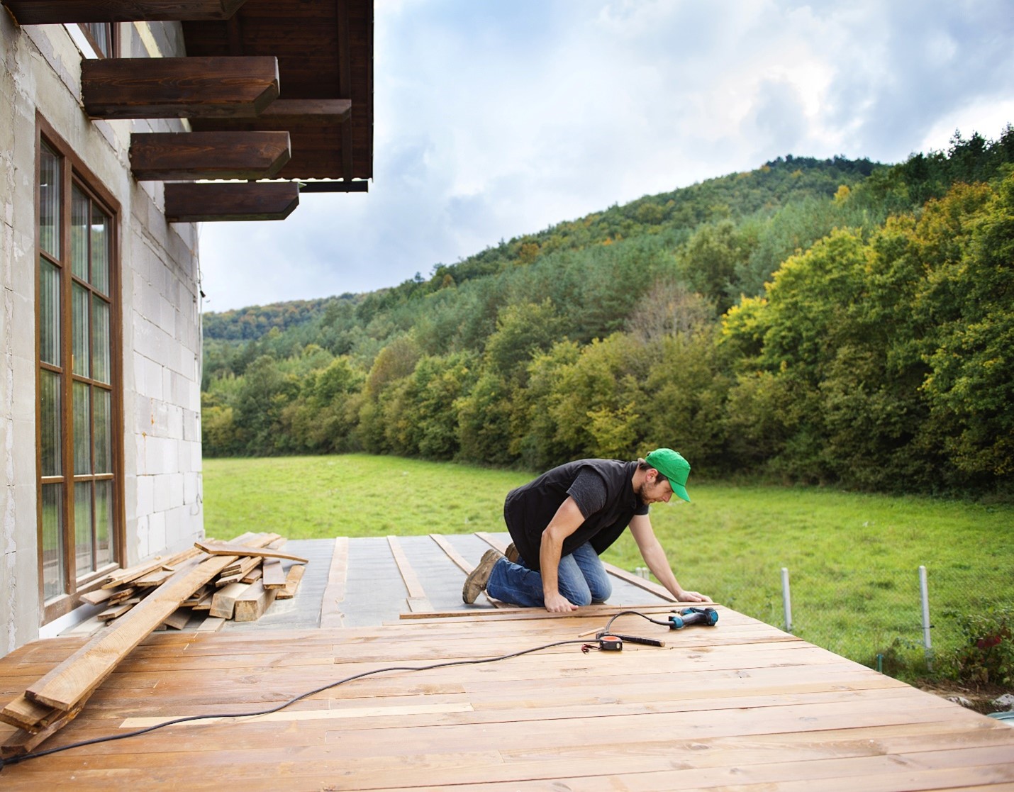 A man focuses on lining up boards for a deck renovation on a home., Behind him are stretching mountains.