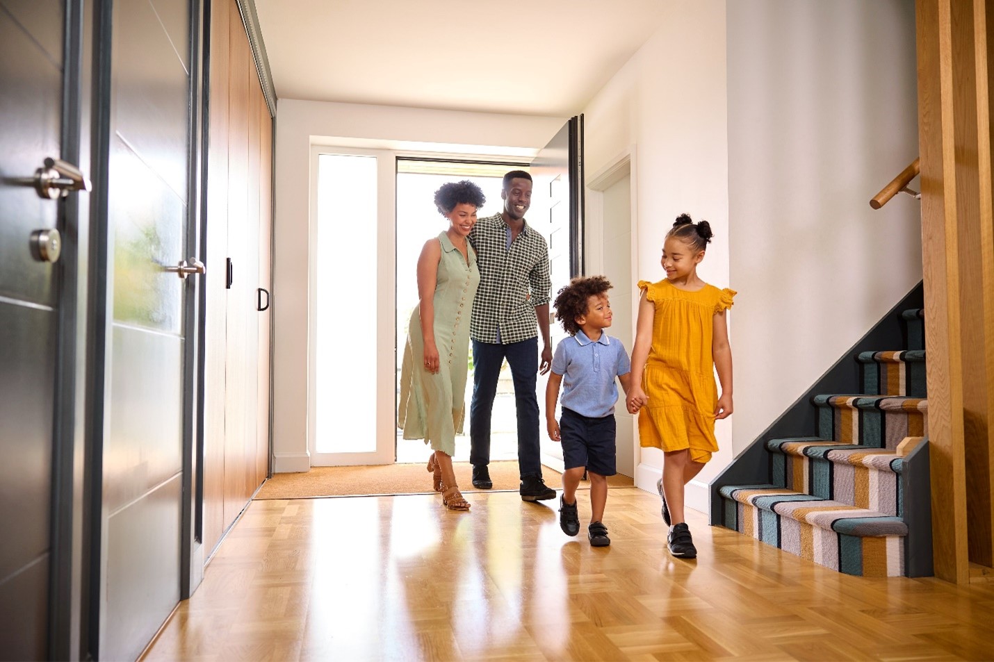 A Black family entering their home. The young brother is holding his big sister's hand Behind them, their father has an arm around their mother. 
