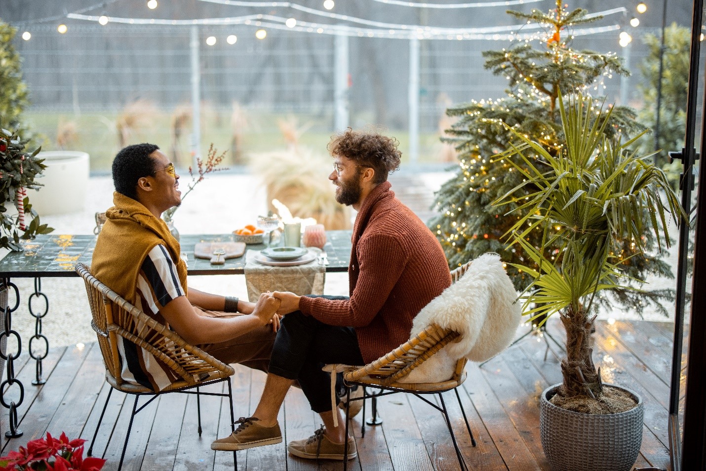 Two masculine presenting people sitting on their porch together, holding hands. They're both smiling and engaged with a conversation together.