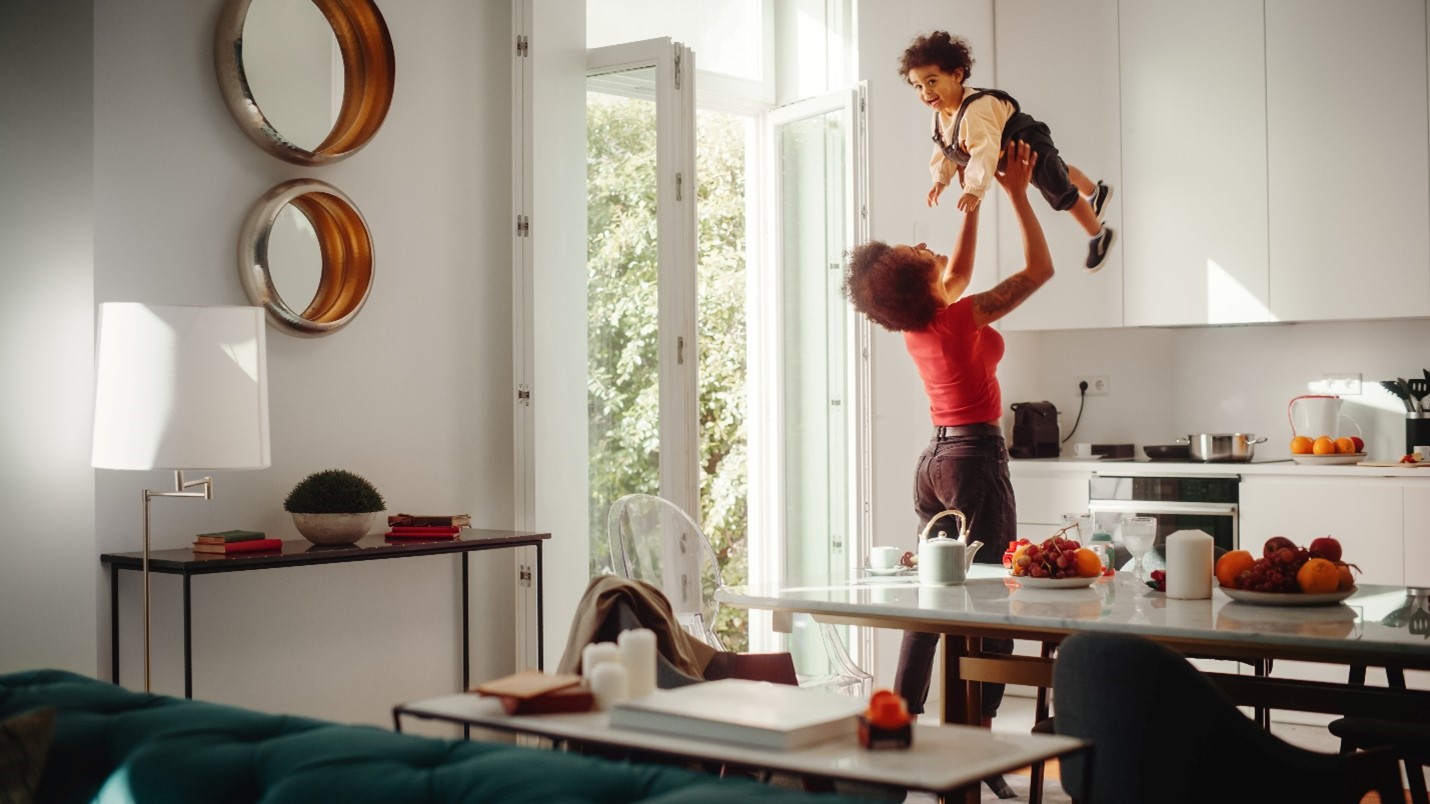 A black mom happily lifting her toddler son into the air in the kitchen of their home. The windows are wide open and various fruits are available on the table.