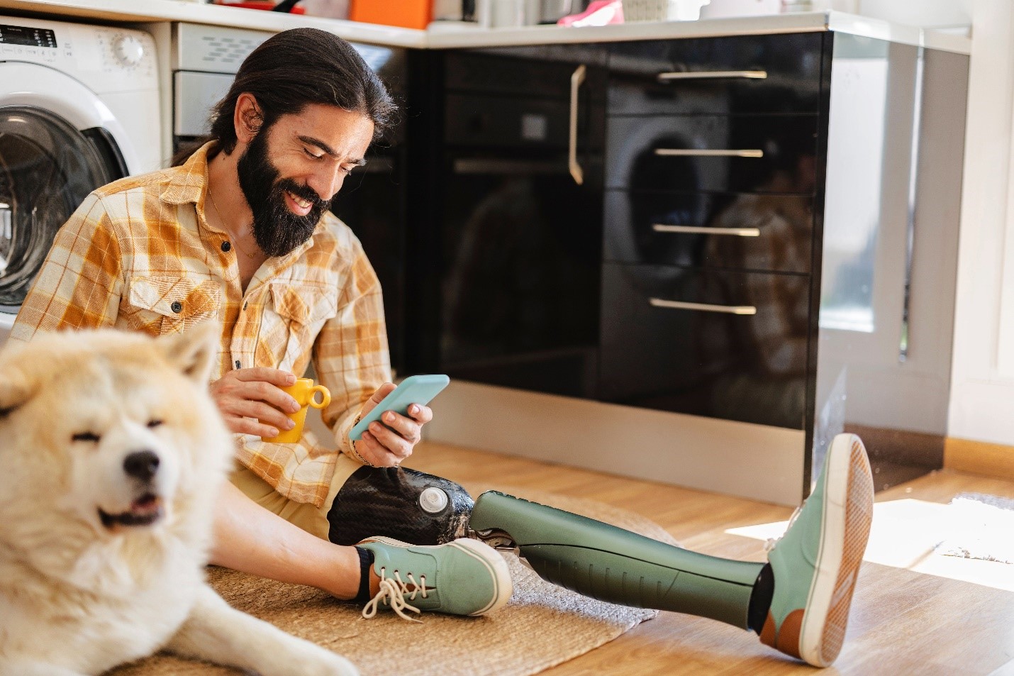 A masculine presenting person with a prosthetic leg sits on the floor of their home next to their dog. They are smiling down at a cell phone in their hand as they hold a cup of coffee in their other.